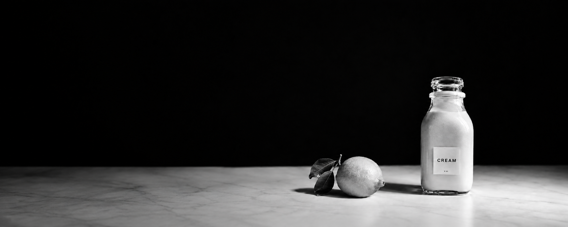 A lemon and a glass bottle of cream arranged on a stone counter in dramatic black-and-white light